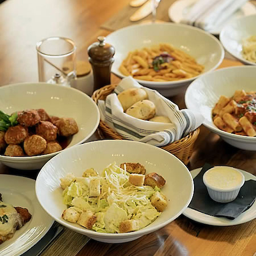 Assorted dishes including pasta, meatballs, salad with croutons, bread rolls, and a small bowl of sauce on a wooden table.