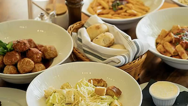 Assorted dishes including pasta, meatballs, salad with croutons, bread rolls, and a small bowl of sauce on a wooden table.