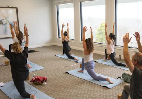 A group of people in a brightly lit room practicing yoga, performing a lunge pose with arms raised on mats.