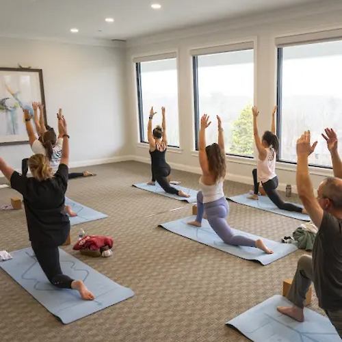 A group of people in a brightly lit room practicing yoga, performing a lunge pose with arms raised on mats.