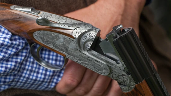 A person holds an intricately engraved shotgun, showcasing its open double barrels against a blue checkered shirt background.