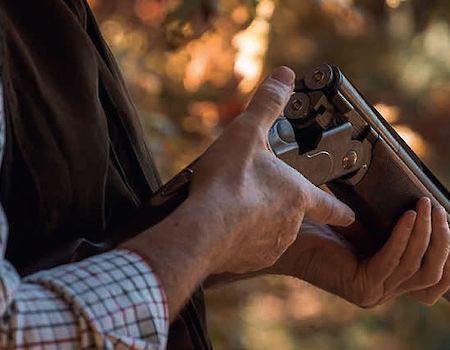 A person is holding and inspecting a shotgun outdoors, wearing a checkered shirt and vest with a blurred natural background in the image.