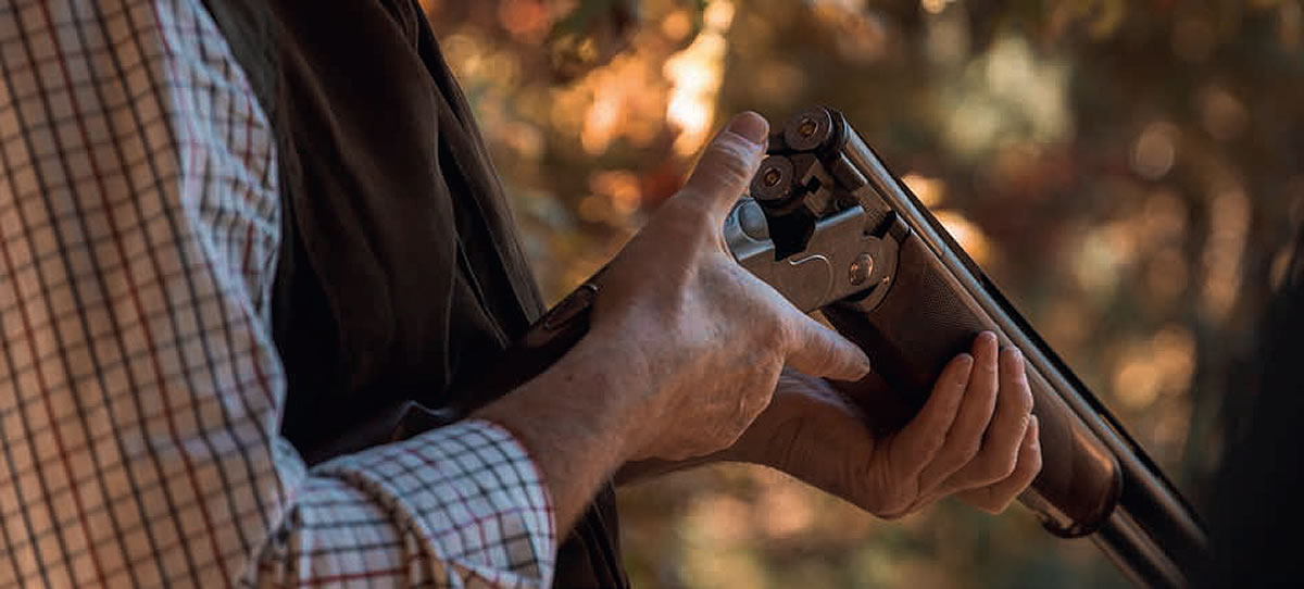 A person is holding and inspecting a shotgun outdoors, wearing a checkered shirt and vest with a blurred natural background in the image.