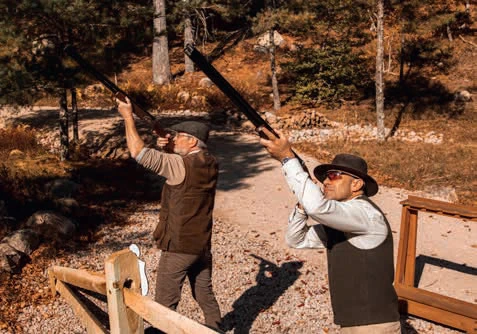 Two people wearing vests and hats are aiming shotguns upward in an outdoor setting.