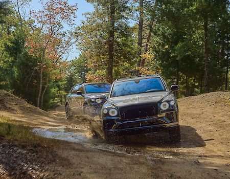 Two SUVs are driving on a muddy, wooded trail, splashing through a puddle surrounded by trees and greenery.