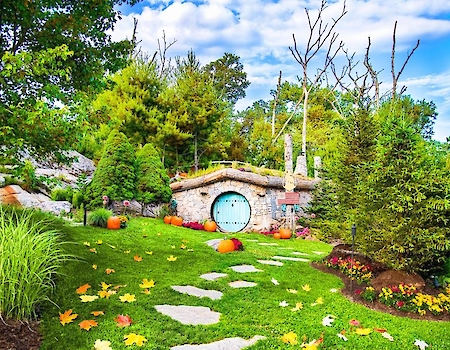 A whimsical garden scene features a round door nestled in a grassy hill, surrounded by trees and autumn leaves, evoking a fairy-tale vibe.