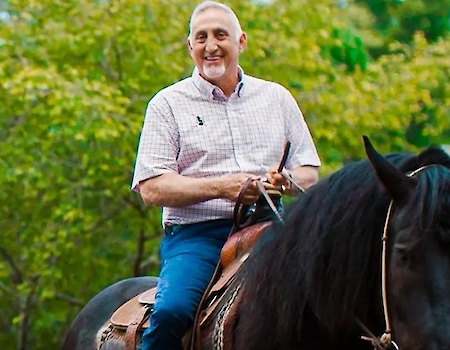 A man is happily riding a dark horse with lush greenery in the background, showcasing a serene outdoor scene.