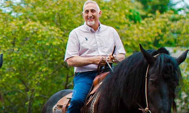 A man is happily riding a dark horse with lush greenery in the background, showcasing a serene outdoor scene.