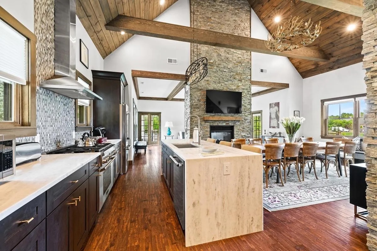 A modern kitchen and dining area with wooden floors, stone accents, high ceilings, and natural light.