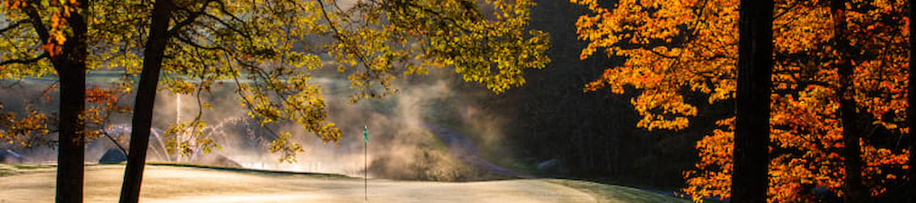 A golf course with autumn trees and morning sunlight filtering through, casting a warm glow and slight mist over the landscape.