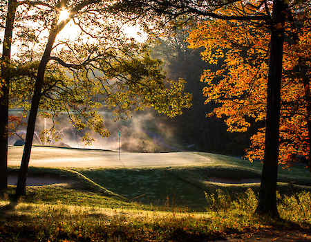 A golf course with autumn trees and morning sunlight filtering through, casting a warm glow and slight mist over the landscape.