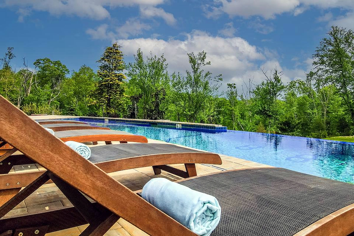 Lounge chairs with towels overlook an infinity pool surrounded by trees under a partly cloudy sky.