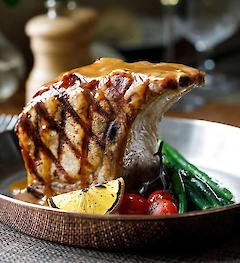 Grilled rib chop with sauce, charred lemon slices, cherry tomatoes, and green beans in a metal pan, served elegantly.