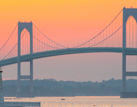 A lighthouse stands before a large suspension bridge against a vibrant sunset sky over calm waters.