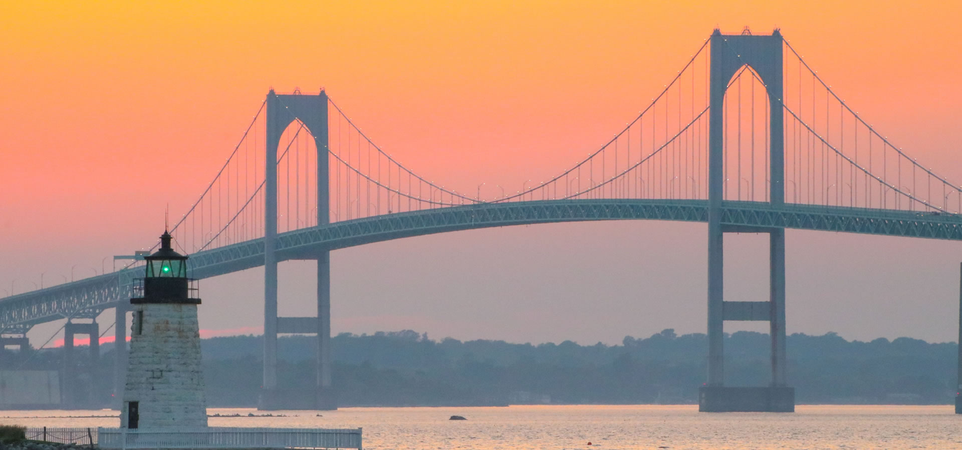 A lighthouse stands before a large suspension bridge against a vibrant sunset sky over calm waters.