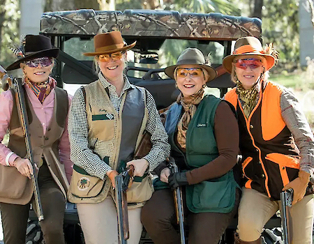 Four people in shooting attire holding shotguns, posing outdoors in front of an ATV with trees in the background.