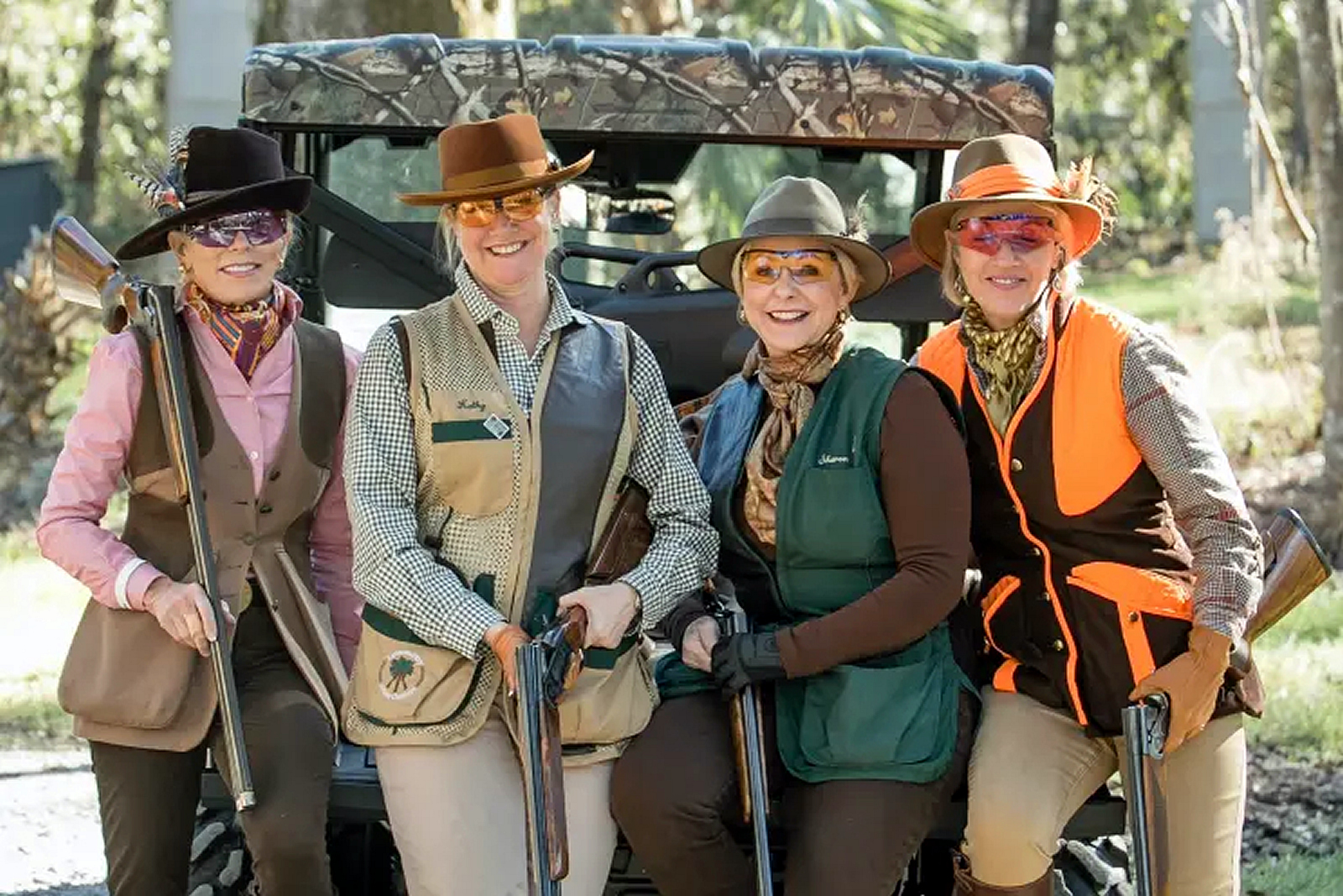 Four people in shooting attire holding shotguns, posing outdoors in front of an ATV with trees in the background.