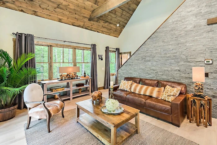 A cozy living room features a leather sofa, wooden coffee table, plants, and large windows with views of greenery outside.