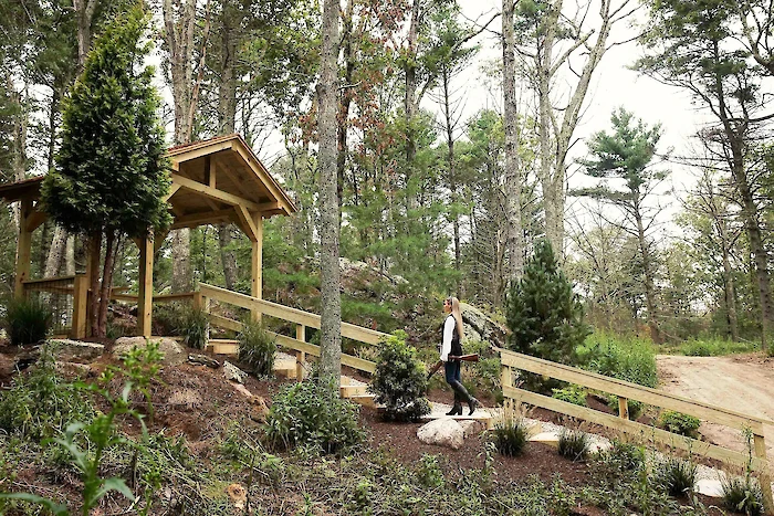 A person walks a dog on a pathway in a wooded area with a small wooden shelter nearby.