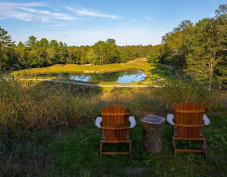Two wooden chairs face a scenic pond surrounded by trees, with a log serving as a side table, under a clear blue sky.