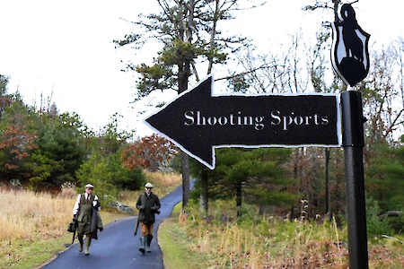 A sign with an arrow reads "Shooting Sports" in a wooded area, with two people walking on a path in the background.