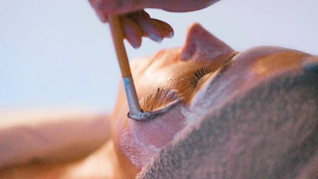 A person is receiving a facial treatment with a brush applying a facial mask while lying down, partially covered by a towel.