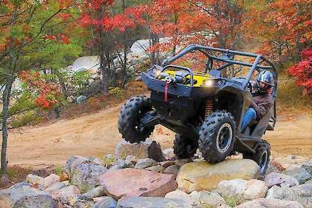 A person driving an off-road vehicle navigates rocky terrain amid autumn trees in a forest setting.