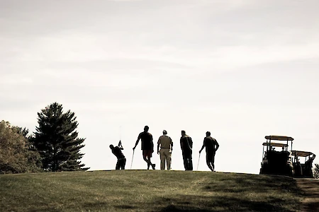 A group of people with golf clubs standing on a hill near a golf cart, silhouetted against a clear sky.