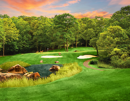 A scenic golf course with a small pond, sand traps, and lush green surroundings under a vibrant sunset sky, surrounded by trees.