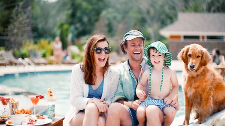 A group of people and a dog are sitting together by a pool, smiling. There's food and drinks on a table nearby.