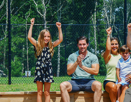 A group of people of various ages are sitting and standing in a bocce court, celebrating with raised arms.