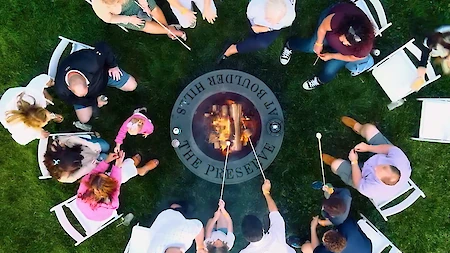 A group of people is seated around a fire pit, roasting marshmallows outdoors. The pit reads "The Preserve at Boulder Hills."