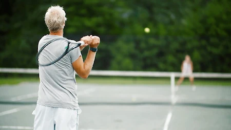 A person in a gray shirt is playing tennis, swinging a racket. Another player is seen on the opposite side of the court.