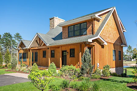 A wooden house with stone accents, surrounded by greenery under a clear blue sky. The design features large windows and a gabled roof.