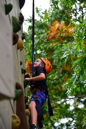 A child wearing a helmet is climbing a rock wall, with a safety harness, surrounded by green and orange foliage in the background.