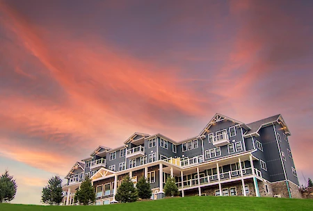 A multi-story building with a dark facade and white trim stands beneath a vibrant, colorful sky at sunset, with trees near its base.