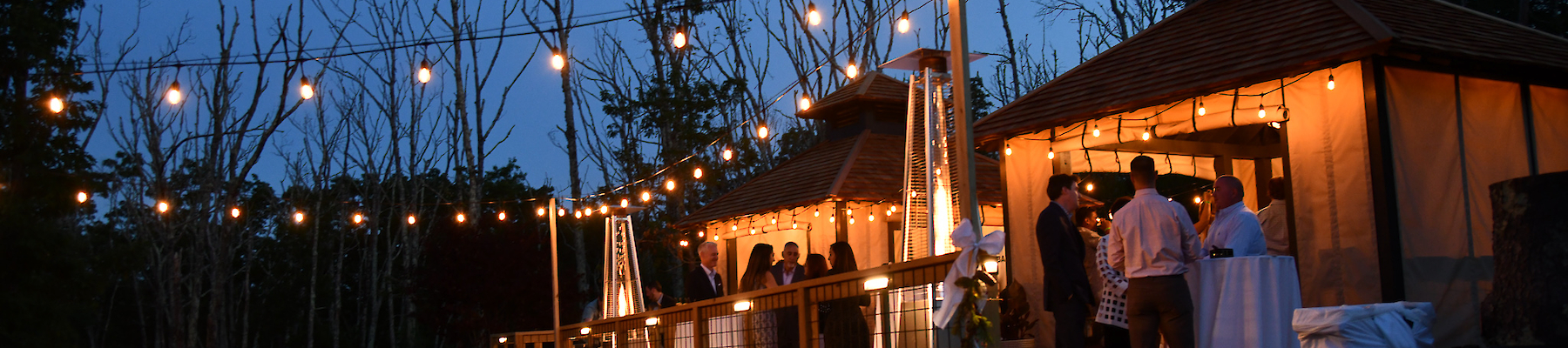 An outdoor event with string lights, a bonfire, and people gathered around a lit-up building in the evening.