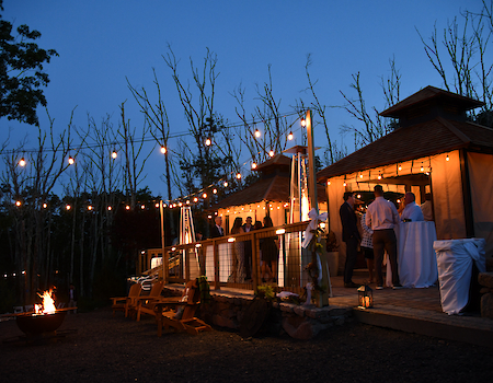 An outdoor event with string lights, a bonfire, and people gathered around a lit-up building in the evening.