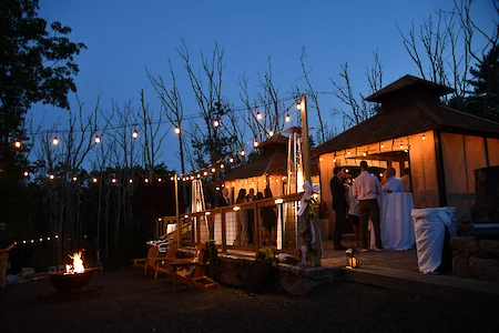 An outdoor evening gathering with string lights, a fire pit, and people socializing near a building.
