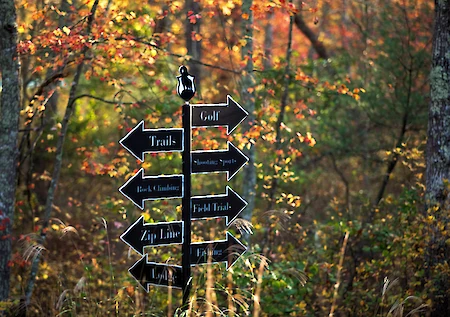 A signpost in a forest with arrows pointing to "Golf," "Trails," "Zip Line," and more, surrounded by autumn foliage.