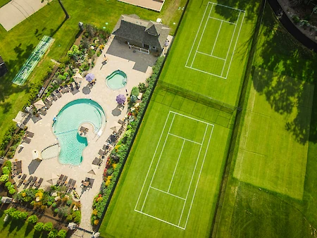 The image shows a swimming pool area with loungers beside two green tennis courts, surrounded by gardens and a small building.
