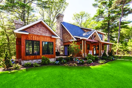 A cozy house with a stone chimney and wooden siding sits amid lush greenery and trees, featuring a well-maintained garden in front.