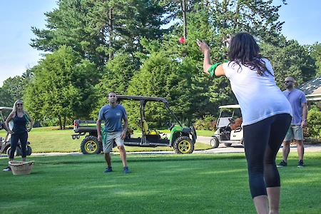 People are playing an outdoor game on a grassy field, with golf carts in the background, surrounded by trees.