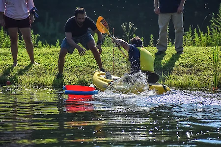 A person in a yellow life vest is paddling a kayak near the shore, creating splashes, while people stand nearby watching.