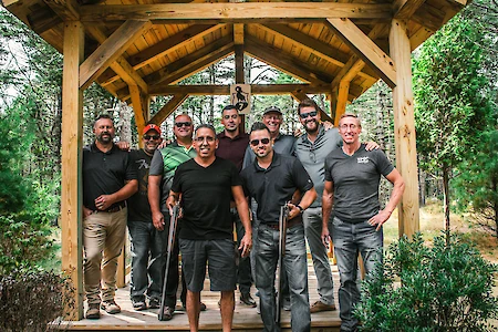 A group of nine men stands under a wooden gazebo in a wooded area, with several holding rifles, dressed casually.
