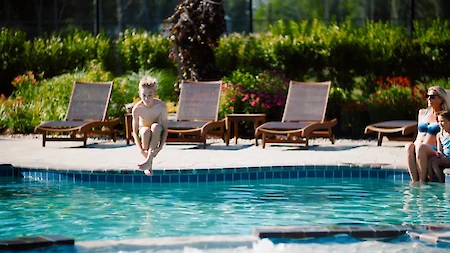 A person jumps into a pool while others relax on lounge chairs beside it.