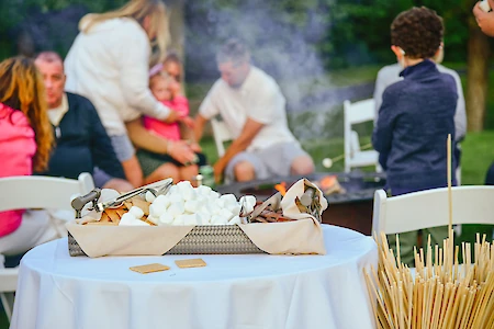 A table with marshmallows, chocolate, and skewers is set up for a bonfire. People are gathered around a fire in the background.