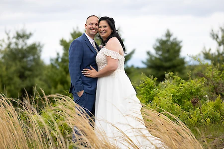 A couple in wedding attire stands smiling in a grassy, outdoor setting with greenery and a cloudy sky in the background.