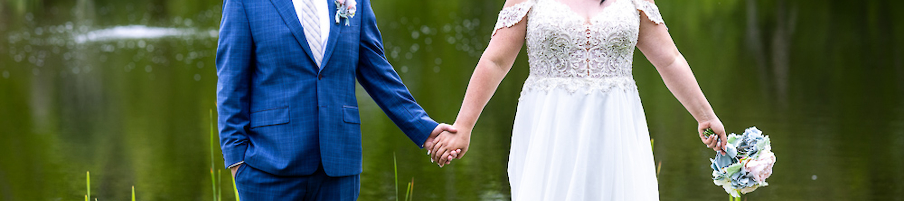 A couple in wedding attire is holding hands by a pond, surrounded by greenery and smiling at each other.