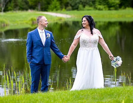 A couple in wedding attire is holding hands by a pond, surrounded by greenery and smiling at each other.